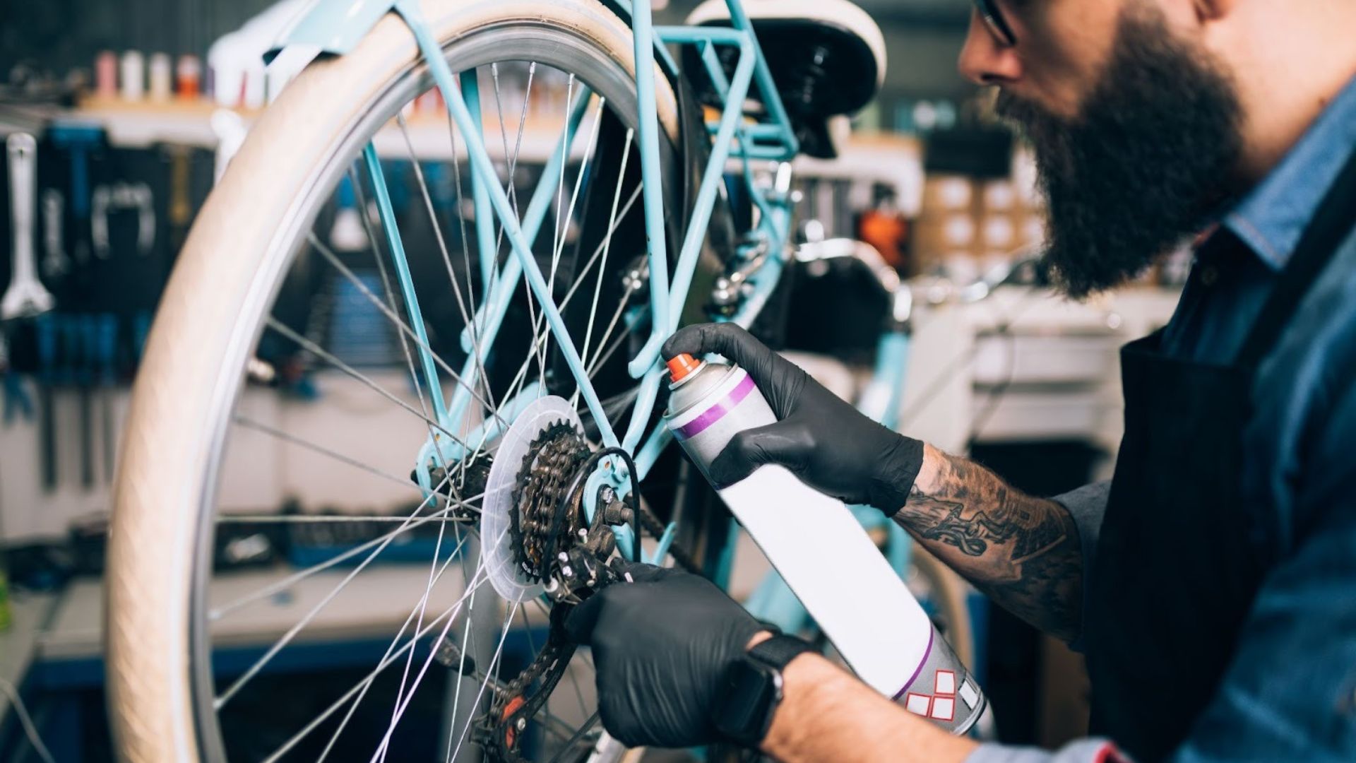 Mechanic wearing gloves applying lubricant spray to the rear gears and chain of a bicycle in a repair workshop